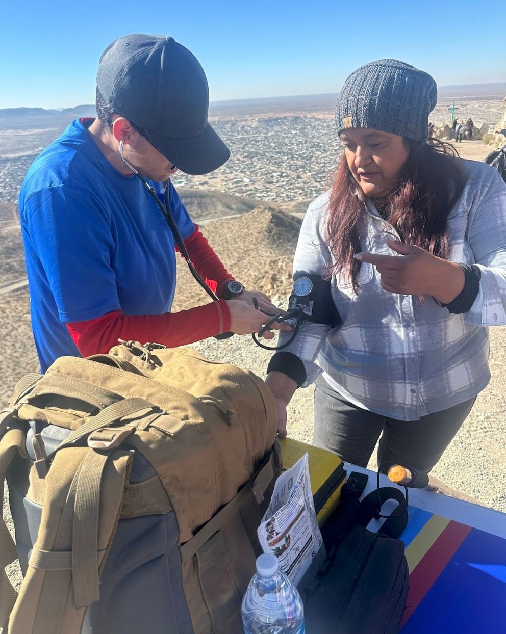 A volunteer in a blue shirt takes a woman's blood pressure at a mountain overlook, a supply pack in front