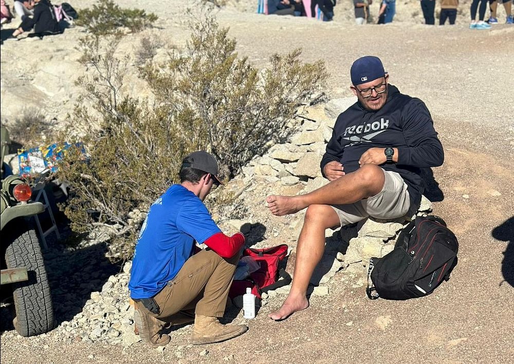 A volunteer in a blue shirt tending a seated man's foot on a rocky hillside