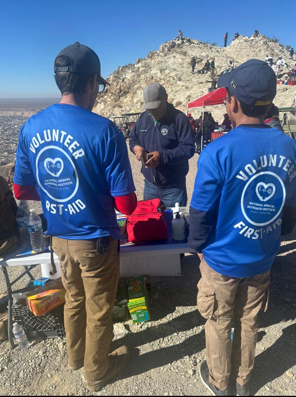 Two volunteers in blue NMOI first-aid shirts stand at a supply table at a mountain event