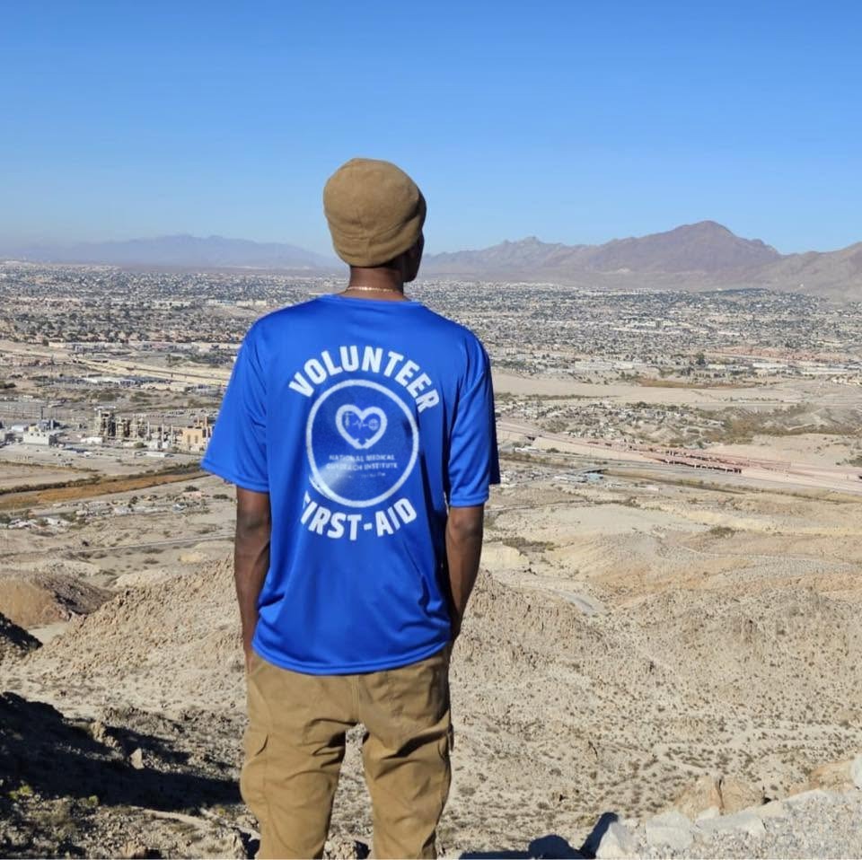 A volunteer in a blue first-aid shirt, seen from behind, looks out over El Paso and the mountains from a ridge