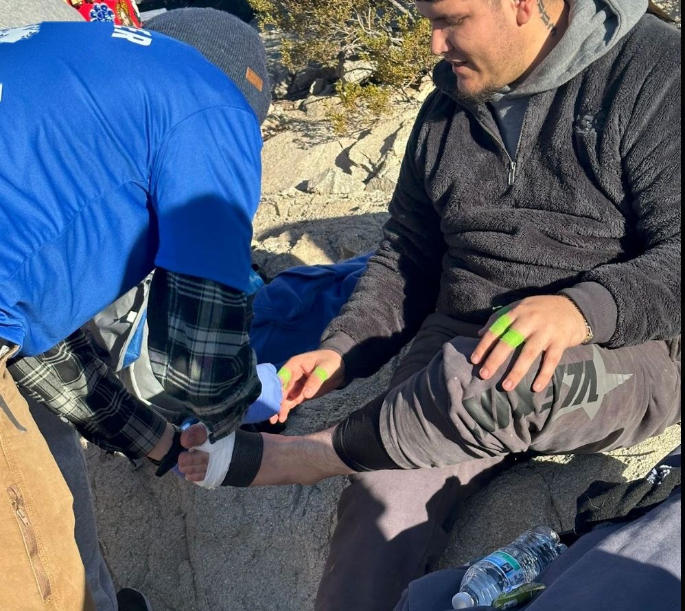 A volunteer wrapping a seated man's foot among rocks